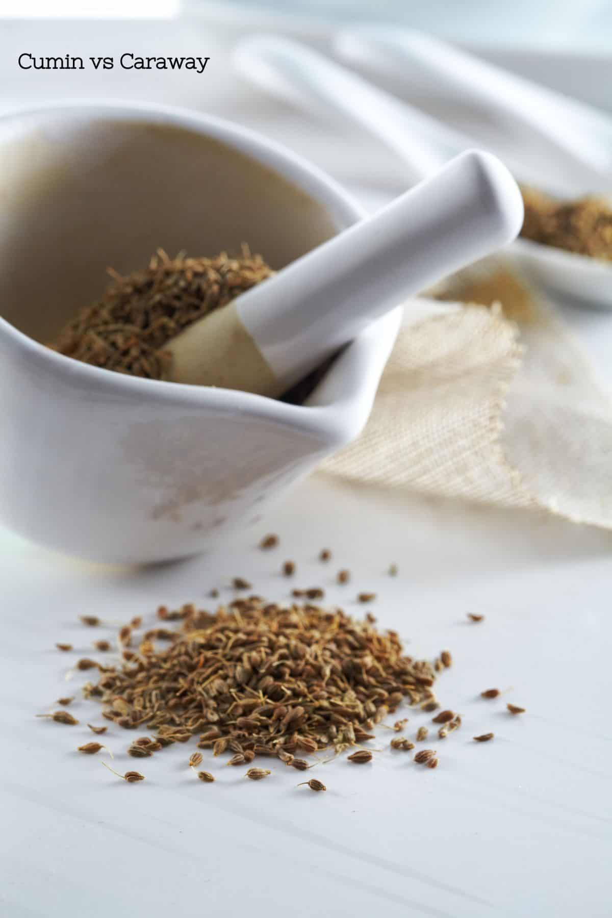 A white mortar and pestle with caraway or cumin seeds in it and in a pile in front of it.