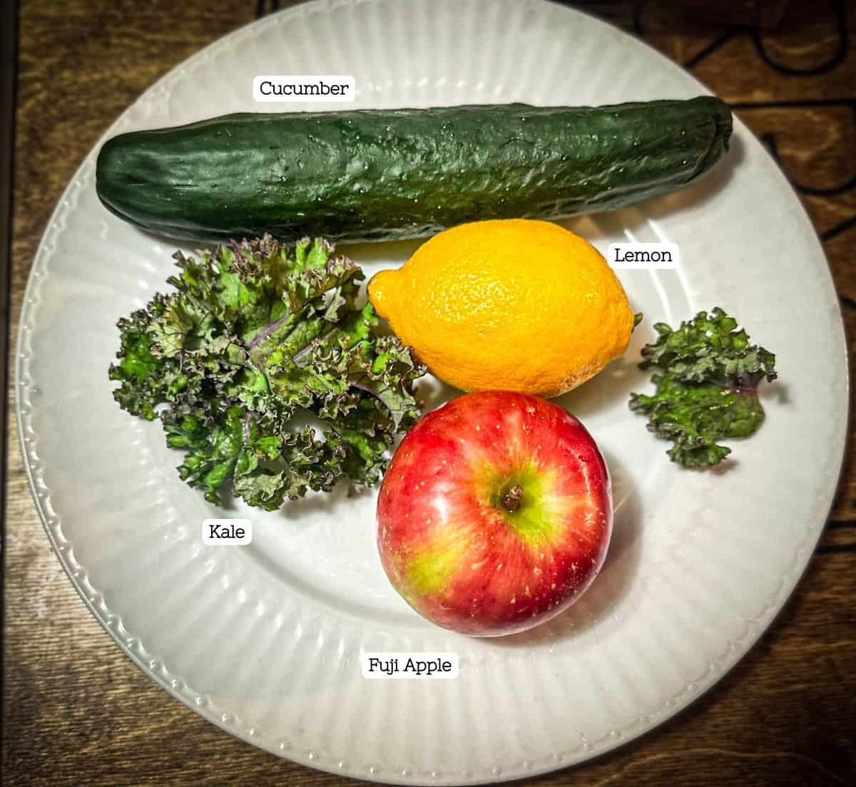 A photo of a white plate with all of the ingredients for first watch kale tonic. There is a purple and green leaf of kale, a yellow lemon, a fuji apple, and an English cucumber at the top.