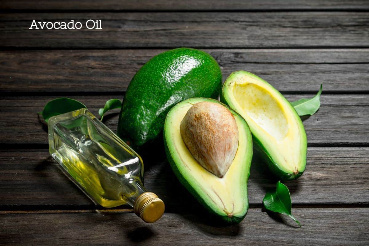 A dark wood table with an avocado and two avocado halves next to a square glass bottle of avocado oil laying on its side.