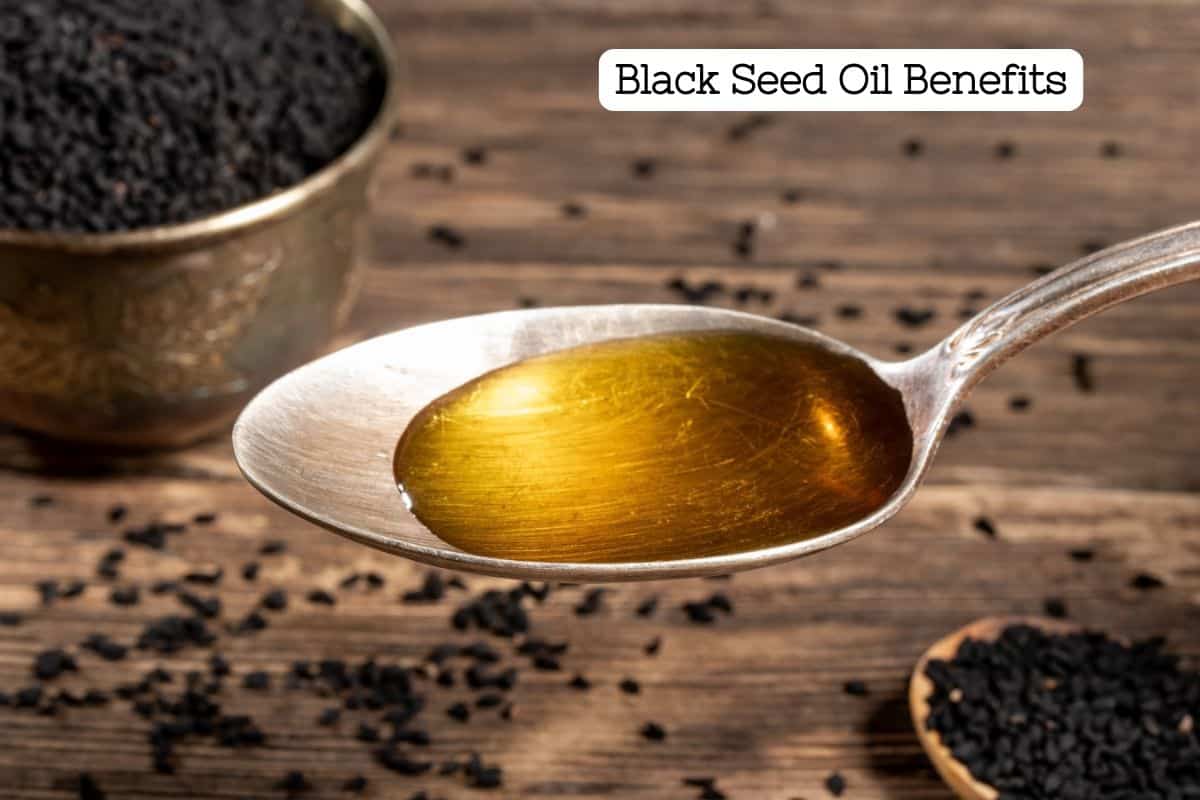 A spoon with golden black seed oil in it and black seeds scattered on a table in the background.