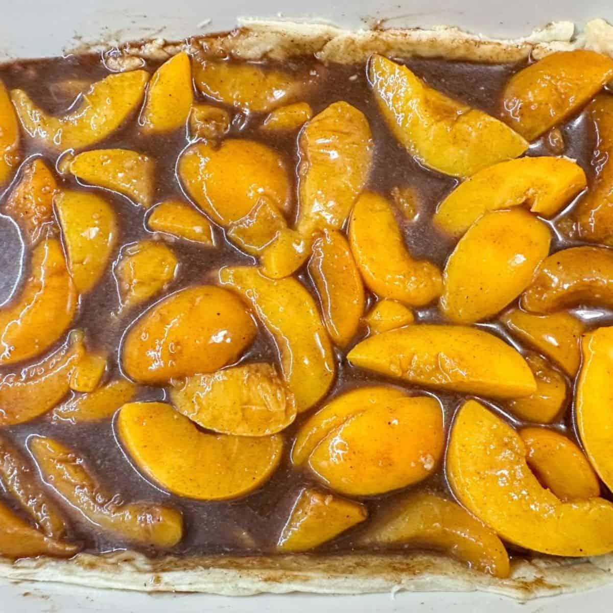 A baking dish with pie crust filled with sliced peaches and thickened brown sauce for peach cobbler.