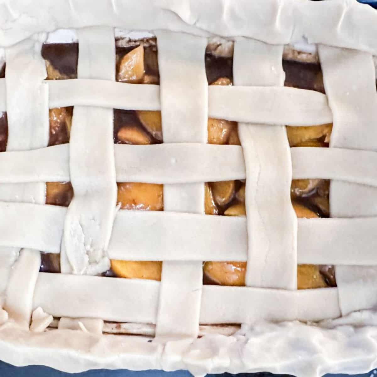 Raw pie crust strips laid atop a peach cobbler in a woven pattern, pre-baking.