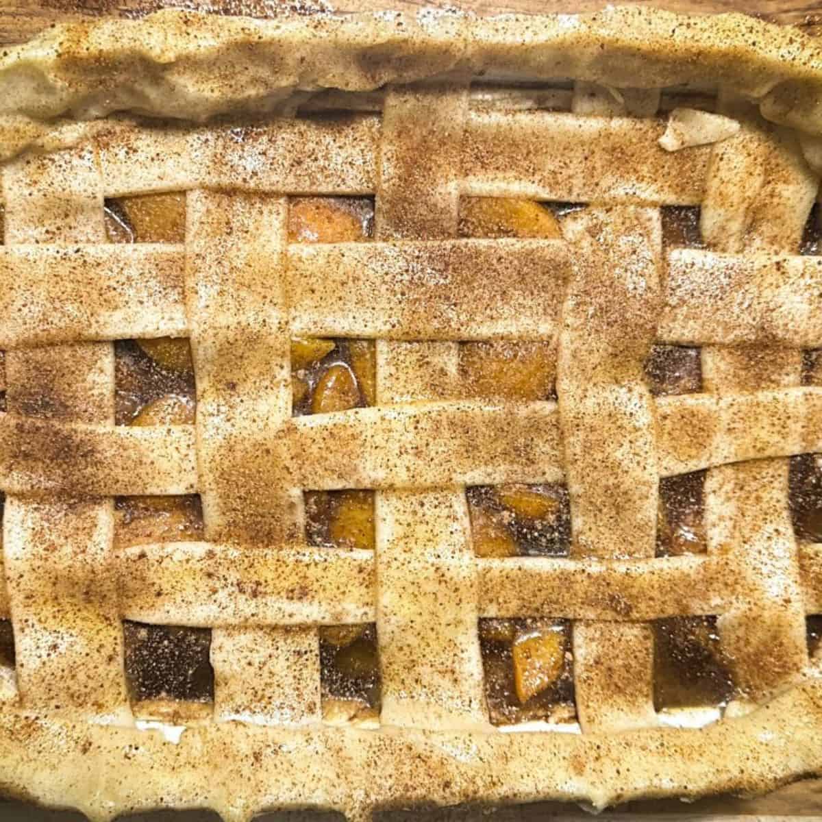 A golden-brown peach cobbler with a woven lattice top, buttered and dusted with spices, ready to bake.