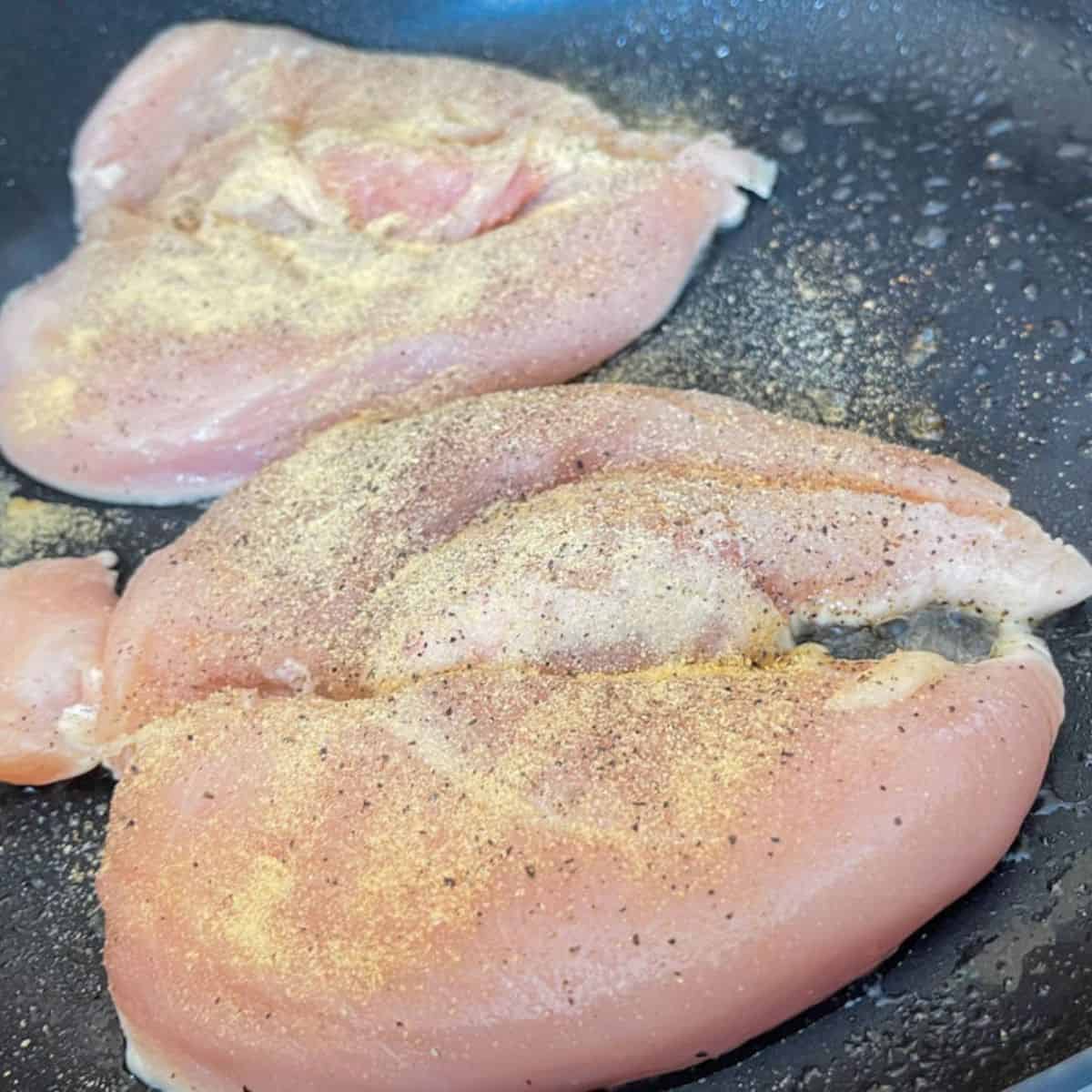 Raw chicken breast butterfly cut and seasoned with onion and garlic powder, salt, and pepper in a skillet seasoned with oil.