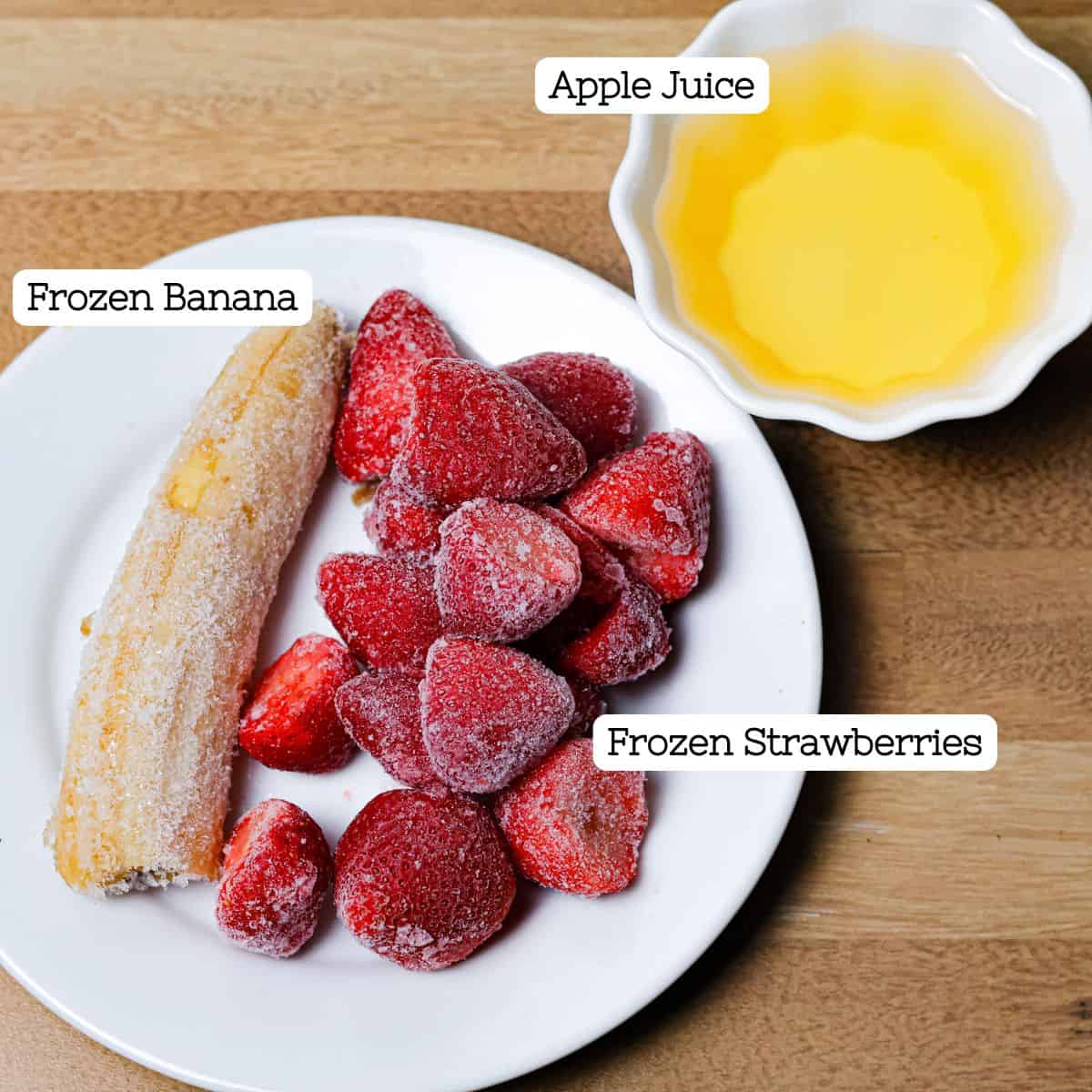 A plate with a whole frozen banana beside a mound of frozen strawberries, with a small bowl of apple juice to the side, ingredients prepped for a smoothie.