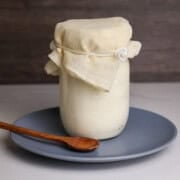Raw Kefir fermenting in a mason jar covered with a cheese cloth on a plate with a wooden spoon.