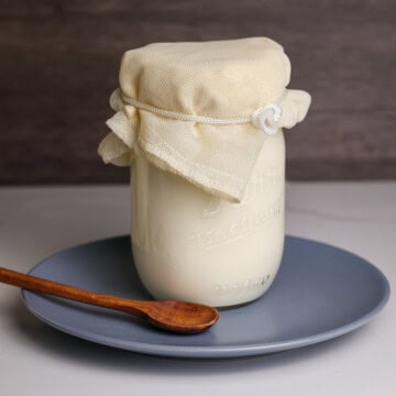 Raw Kefir fermenting in a mason jar covered with a cheese cloth on a plate with a wooden spoon.