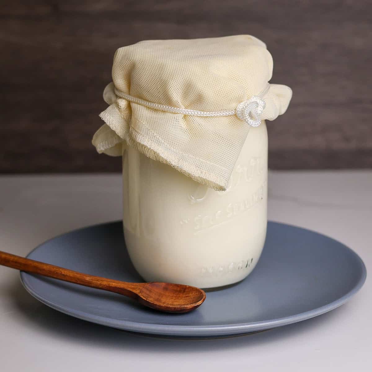 Raw Kefir fermenting in a mason jar covered with a cheese cloth on a plate with a wooden spoon.