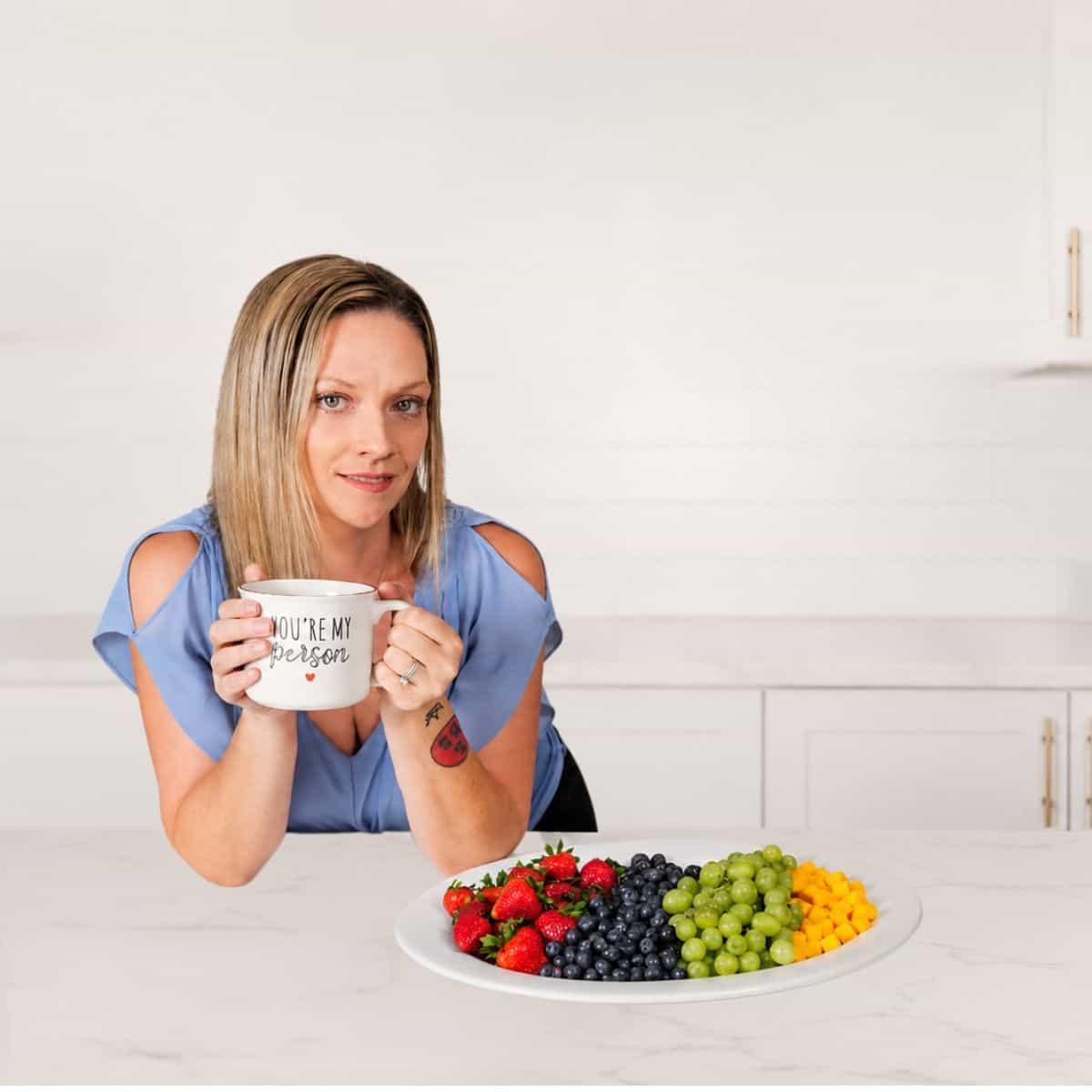Natalie holding a coffee mug in a kitchen next to a plate of fresh fruit.